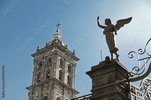 Stone angel statue welcoming visitors at entrance with majestic bell tower of Cathedral rising behind under clear blue sky in Puebla, Mexico