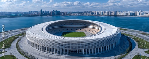 Aerial view of a modern circular football stadium located on a coastal waterfront area with a city skyline under a blue sky, concept for sports infrastructure, urban development and venue