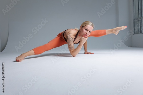 **Stock Photo Description (English):**

Fit athletic woman practicing advanced yoga balance pose on a light studio background. She is wearing a brown top and orange leggings, holding her body in a str
