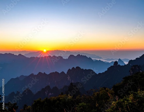 A breathtaking panoramic view captures a vibrant sunrise over a sprawling mountain range, bathed in hues of orange, yellow, and blue, creating a serene and majestic landscape.