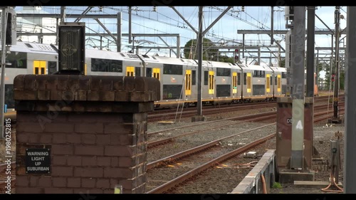 Passenger train moving through an industrial rail corridor in Sydney, Australia, with tracks and infrastructure in the foreground and warehouses in the background. Clean, commercial shot.