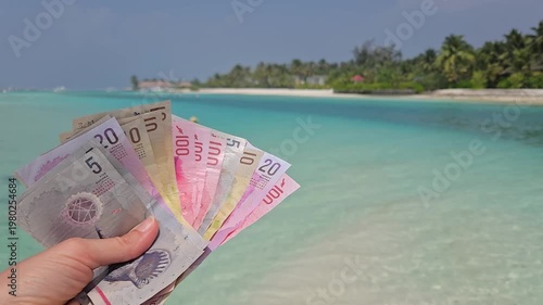 Stack of different denominations of Banknotes of Maldives, Maldivian rufiyaa in hand with maldivian island and ocean in background. Expensive travel concept