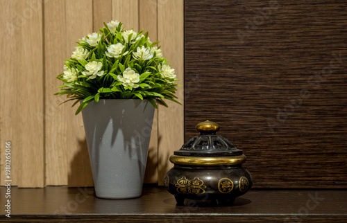 Potted green plant with small white flowers placed beside a decorative incense burner on a wooden shelf, creating a calm and natural interior setting.