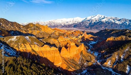 A stunning aerial view showcases colorful canyons and snow-capped mountains under a clear blue sky.
