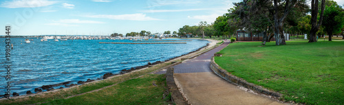 Принти на полотні Waterfront Park Walkway Along Calm Marina With Boats, Trees and Grassy Promenade