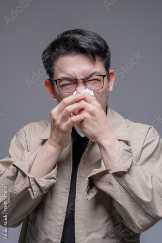 Фотография Asian man with glasses blowing his nose with a tissue, suffering from a cold, allergy, or PM 2