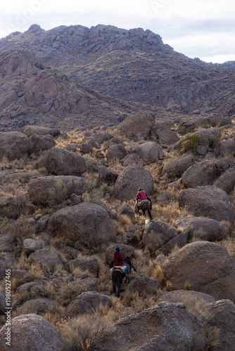 Photos Traditional Argentine gauchos riding horses through a rugged mountain terrain with large granite boulders under a cloudy sky, rural Andean culture