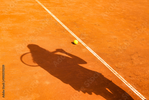 Tennis Match on a Clay Court. A lone tennis ball sits near the baseline, as the long shadow of a player holding a racket stretches across the red clay court on a bright, sunny afternoon.