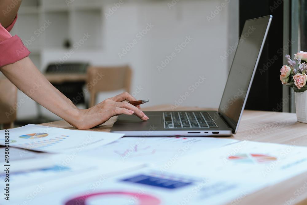 Fototapeta premium A professional reviews financial reports, pointing at charts while working on a laptop, analyzing data and planning business strategy at a modern office desk.