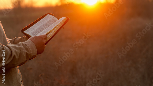 Person reading a Bible outdoors during a warm sunset with golden light illuminating the scene