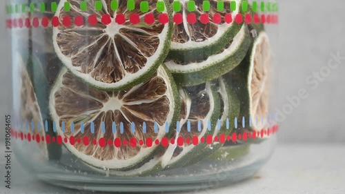 Dried lime slices inside glass jar with red green patterns rotating on white table surface.