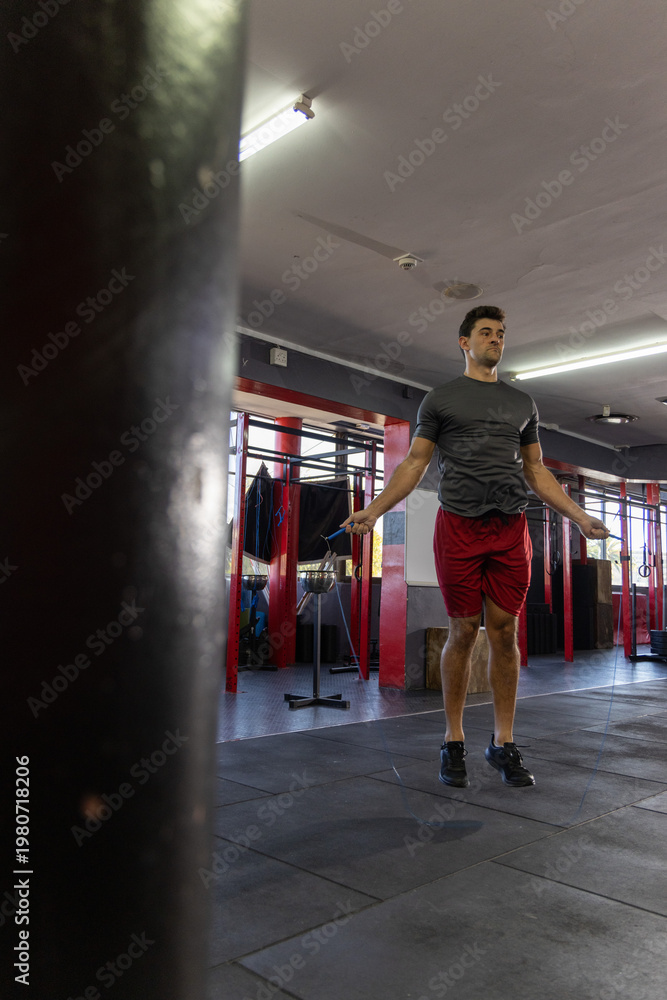 Fototapeta premium Mid-adult man jumping rope in gym near heavy bag, pull-up rig wearing grey t-shirt red shorts