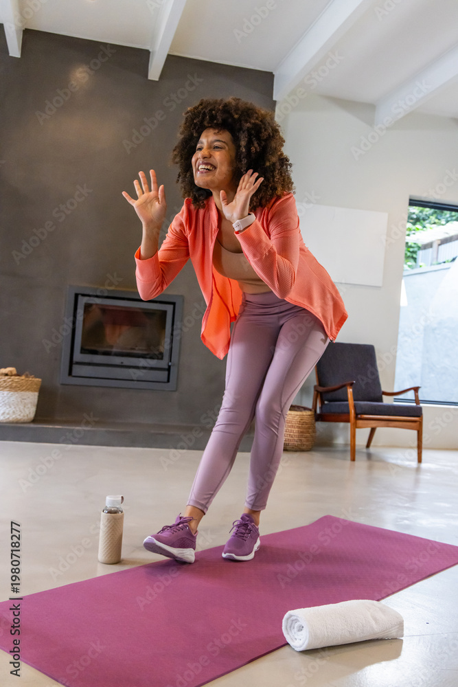 Naklejka premium African American woman balancing on magenta mat in living room wearing coral jacket, wrist tracker