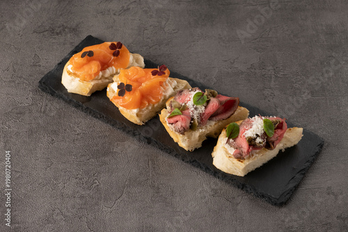 Gourmet canapes featuring smoked salmon, beef, and cream cheese on a black slate platter against a textured gray background