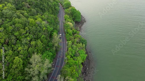 Drone footage reveals lush green forest flanking a winding road along the water. A paved path curves along the tranquil natural edge of the river, creating a beautiful arc.