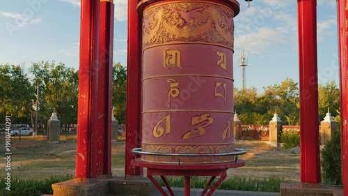 A large red Buddhist prayer wheel with golden mantras spins at sunset. This traditional religious object is bathed in the warm light of the sunset, creating a serene and spiritual atmosphere