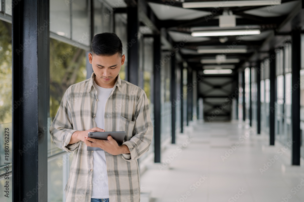 Fototapeta premium Young asian man looking at his tablet in a modern glass hallway. working online. Work from anywhere.