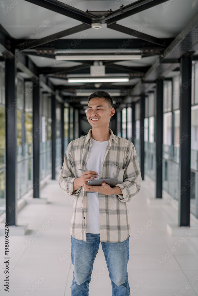Fototapeta premium Smiling asian man standing and look at to the window holding tablet in a modern glass hallway. working online. Work from anywhere. modern tech