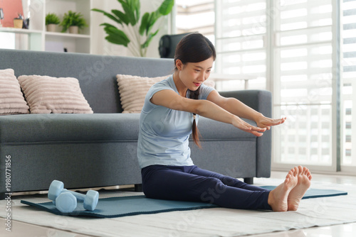 Asian adult woman stretching doing home yoga workout on floor mat with dumbbells in modern apartment living room practicing healthy fitness routine