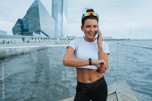 A woman in sporty attire enjoys a phone call by the waterfront, exuding joy and relaxation while embracing the dynamic urban environment around her.