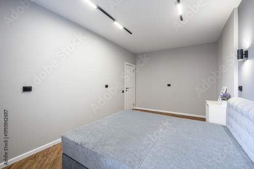 A minimalist bedroom with a grey upholstered bed, light grey walls, wooden flooring, a white nightstand with a vase of lavender, recessed ceiling lights, and a white door.