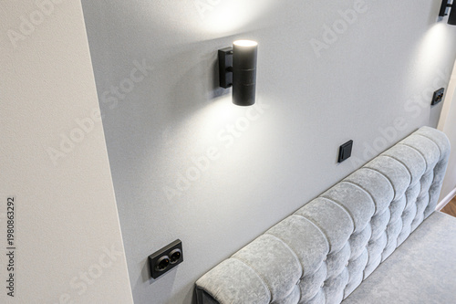 A modern bedroom corner with a grey tufted headboard, a sleek black wall lamp casting soft light, black sockets and switches on a light grey wall.