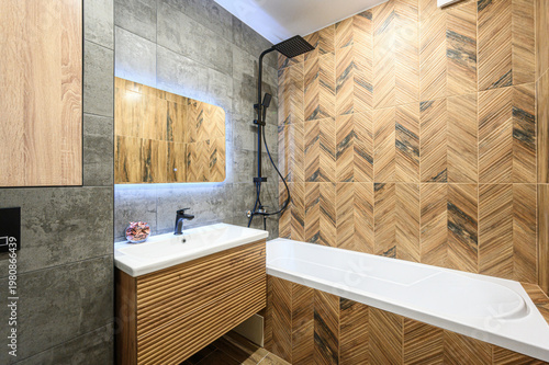 Cuadro en lienzo A modern bathroom with herringbone wood-pattern tiles, grey concrete-style walls, white bathtub, sleek sink with wooden vanity, black showerhead, and backlit mirror