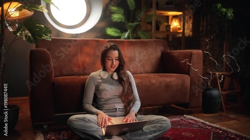 Young woman sitting on floor with laptop and having a video call in a cozy living room