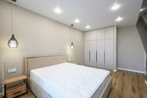 A modern bedroom with a large bed, beige upholstered headboard, wooden nightstand, pendant lights, built-in wardrobe, light wood flooring, neutral walls, recessed ceiling lights.