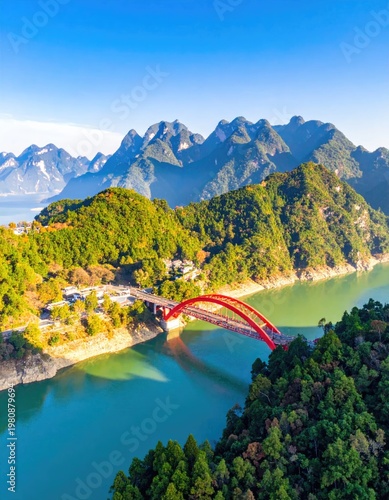 A vibrant aerial view showcases a striking red arch bridge spanning a serene turquoise river, framed by lush green hills and a majestic mountain range under a clear blue sky.