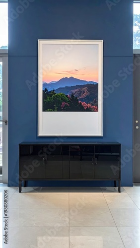 A framed landscape photograph depicting a vibrant sunset over rolling hills and lush greenery is displayed on a blue wall above a sleek black cabinet in a modern interior setting.