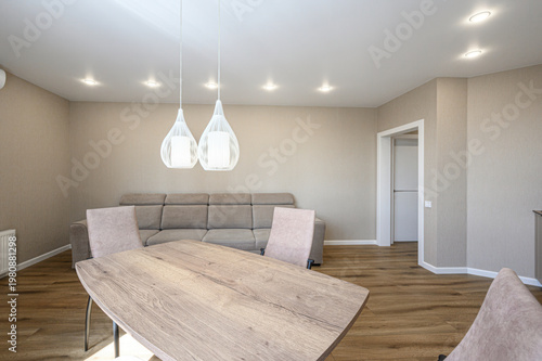 A modern living room with a beige sofa, wooden dining table, matching chairs, pendant lights, recessed ceiling lights, light beige walls, wooden flooring, and a clean, minimalist design.
