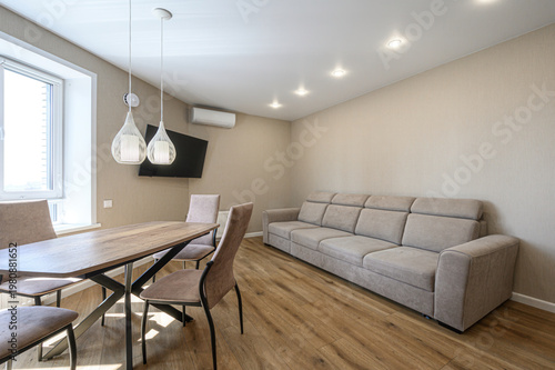 A modern living-dining area with a beige sofa, wooden dining table, pendant lights, wall-mounted TV, air conditioner, light beige walls, wooden flooring  bright and minimalist.