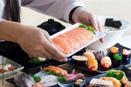 Close-up of hands serving a fresh salmon sashimi platter on a wooden table, surrounded by various Japanese dishes in a restaurant setting.