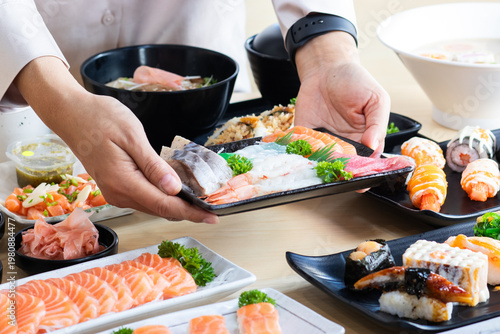 Close-up of a person serving a fresh mixed sashimi platter including salmon, tuna, and shrimp, part of an abundant Japanese meal on a wooden table.