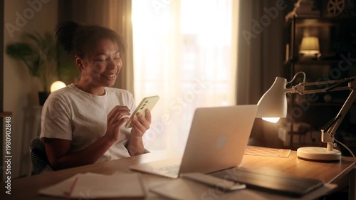 Happy african american woman scrolling on smartphone while sitting at desk with laptop in sunny room