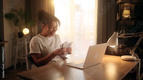 Happy african american woman counting dollar cash banknotes while sitting at desk with laptop