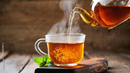 Fotografía Hot tea is poured from a glass teapot into a clear mug with fresh mint on wooden