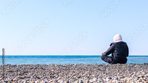 Seated figure in winter clothes on beach staring at ocean