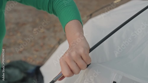 Woman assembling tent poles during camping, closeup of hands fitting collapsible pole segments into sleeve and clip, green sweater and jeans visible, forest campsite with fallen leaves, calm
