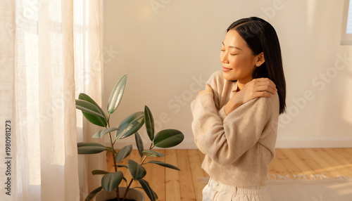 A woman in cozy clothing enjoys a peaceful moment hugging herself beside a large window with sunlight and a green indoor plant nearby.