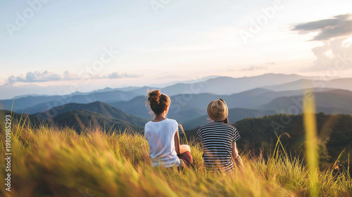 Two people sitting together on a grassy hill, looking at a layered mountain range during a beautiful sunset