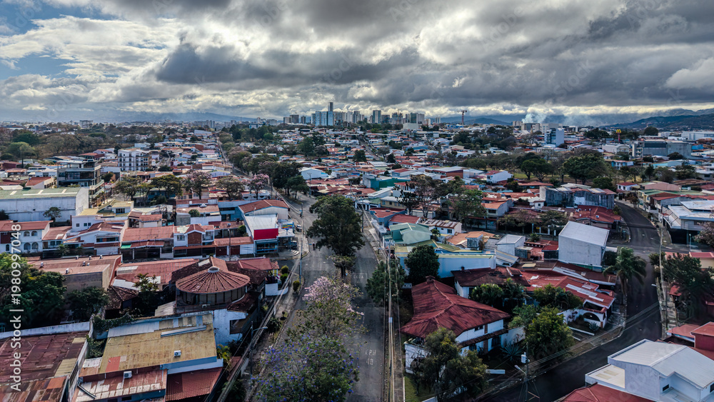 Obraz premium Drone aerial view of downtown San Jose, Costa Rica, showcasing dense urban neighborhoods, city skyline, dramatic cloudy sky, a vibrant Central American capital with mixed architecture (February 2026)