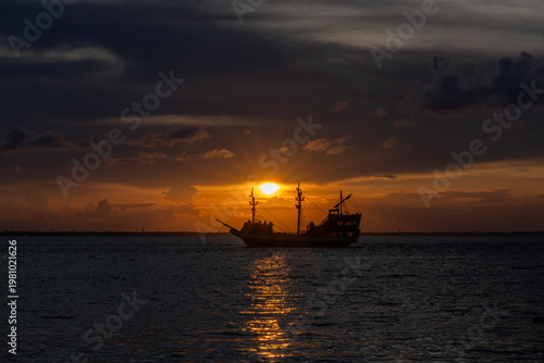 A sailing ship is silhouetted against a vibrant sunset, casting a warm glow over the ocean. The sky's hues range from deep orange to soft pink, reflecting beautifully on the water's surface.