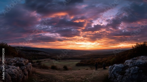 Two figures overlook a sprawling vast landscape beneath a dramatic richly colored sunset sky at dusk