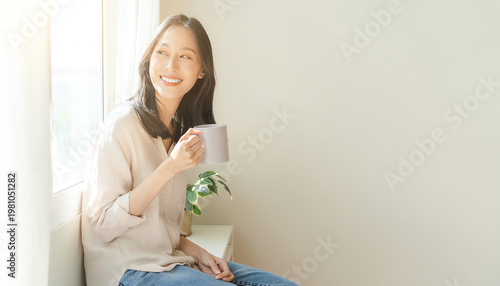 A smiling young woman sits by a bright window, holding a cup, enjoying a peaceful moment indoors with natural light and a small plant nearby.