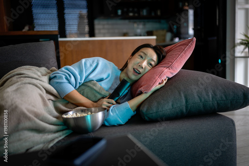 Smiling young woman lying on a sofa with popcorn, watching a movie at night—cozy home entertainment, relaxed mood, and comfortable evening leisure