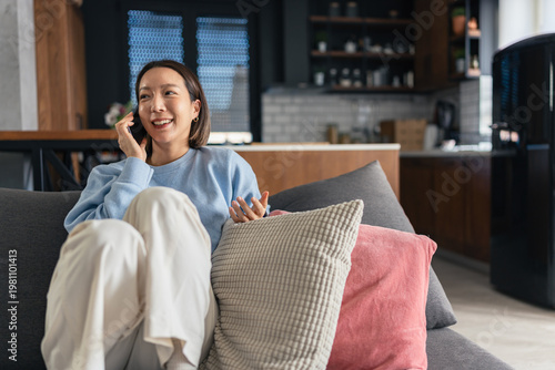 Tableau sur toile Smiling Asian woman talking on smartphone while relaxing on a sofa at home