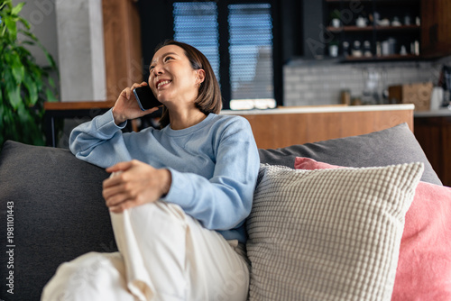 Schilderij op canvas Smiling Asian woman talking on smartphone while relaxing on a sofa at home