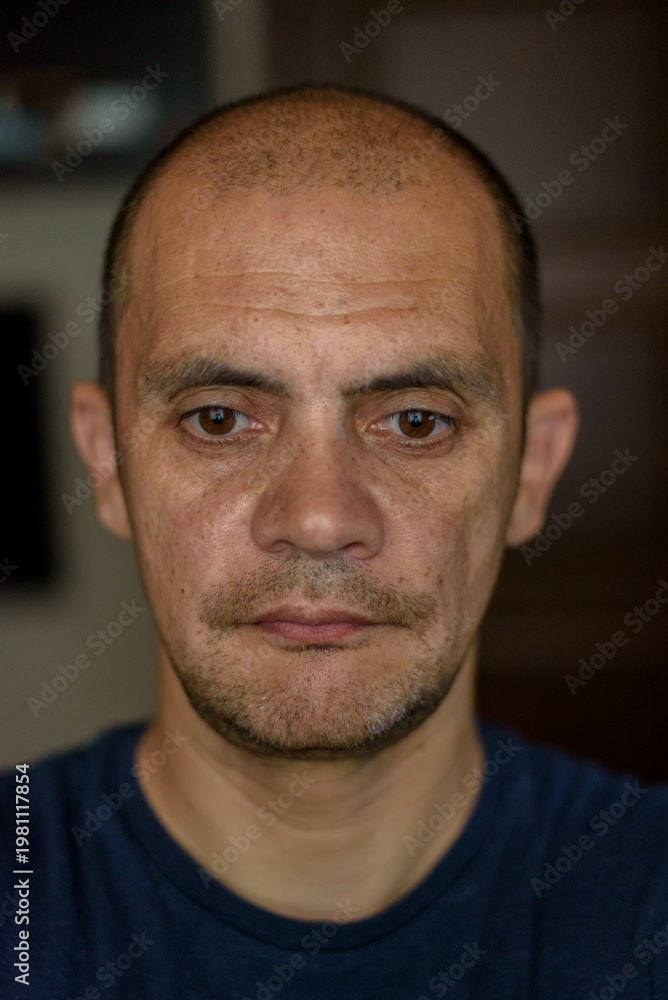 Fototapeta premium Middle-aged man with short hair and facial hair, wearing a blue t-shirt, poses for a close-up portrait against a blurred background
